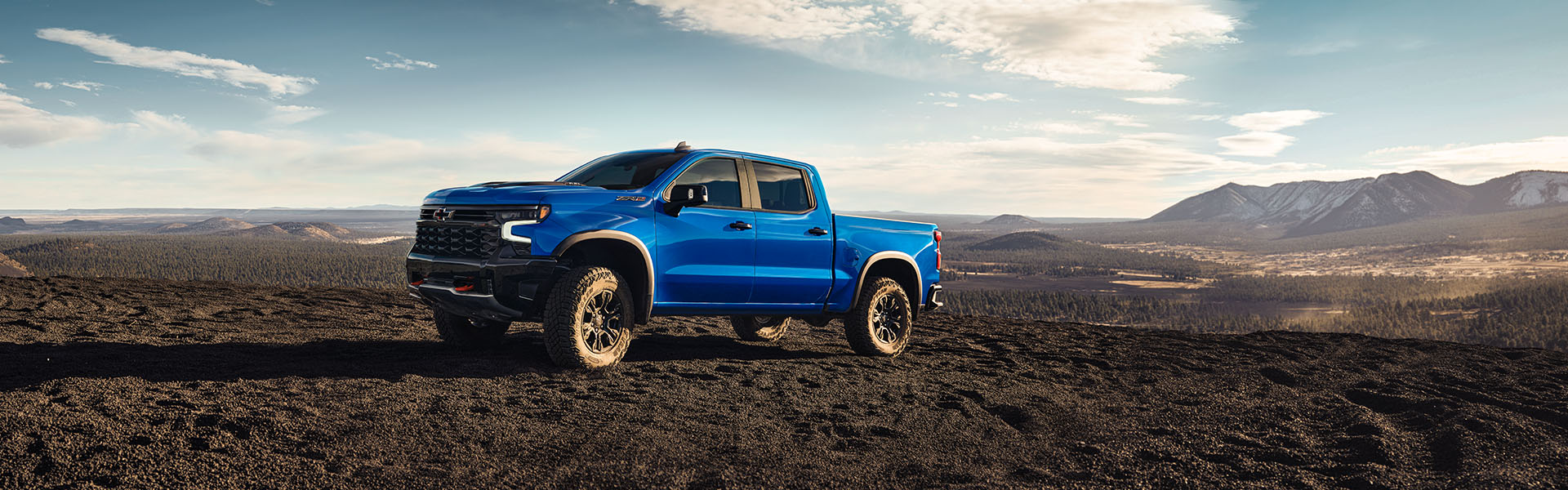 2025 Chevrolet Silverado 1500 in bright blue parked on rugged terrain with mountains and wide-open desert landscape in the background.