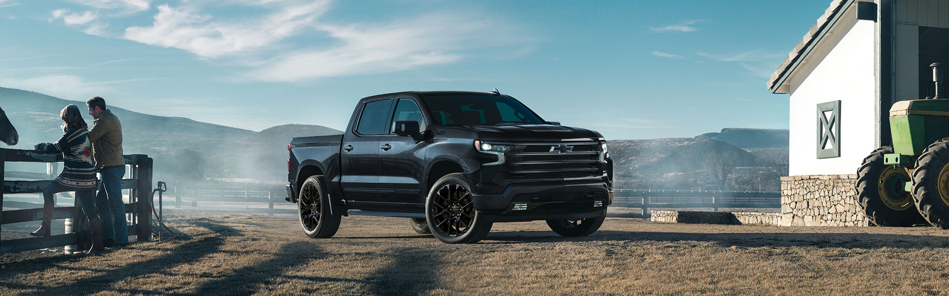 2025 Chevrolet Silverado 1500 in black shown from a front three-quarter angle on a rural farm setting, featuring a crew cab design, and rugged pickup styling with mountains in the background.