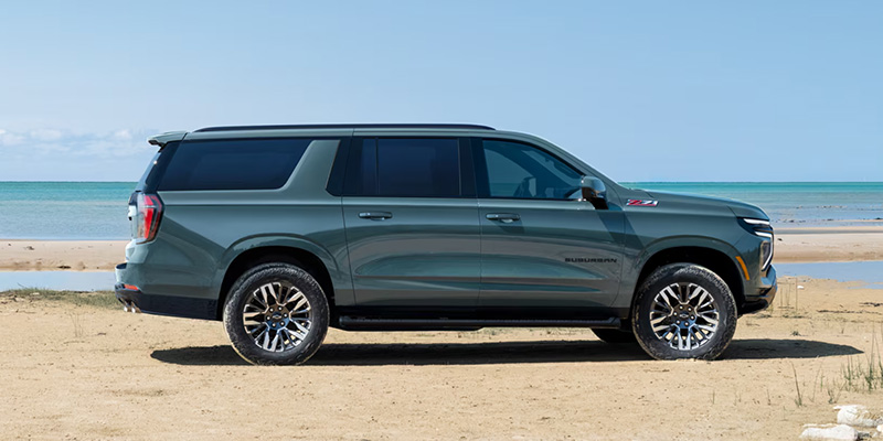 A 2026 Chevrolet Suburban parked on the beach, with waves and sand in the background under a clear blue sky.