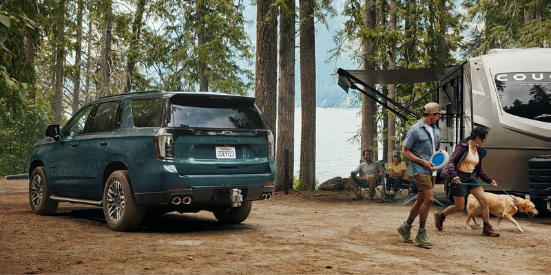 A green Tahoe is parked in a forest campsite near a lake. Two people walk a dog past an RV; others sit nearby. The scene feels adventurous and relaxed.