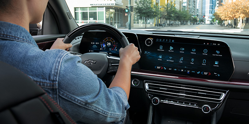 A person drives a modern Traverse in a city, hands on the steering wheel. The dashboard features a large digital touchscreen with various app icons.