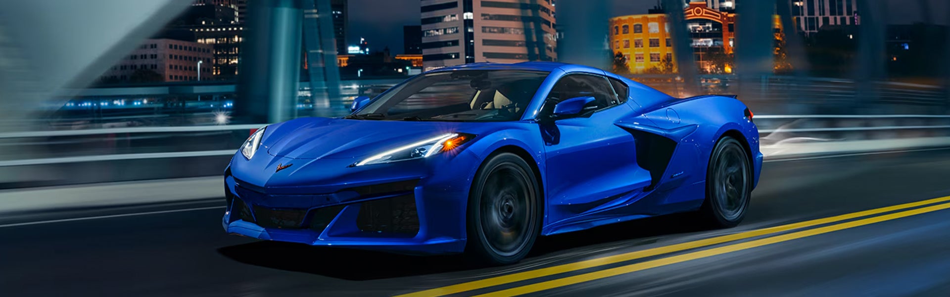 A 2026 Chevrolet Corvette E-Ray driving on a highway at night, illuminated by streetlights and city lights in the background.